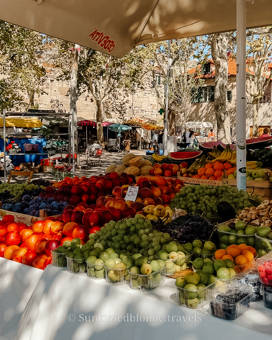 Fresh fruit stall at Split&rsquo;s outdoor Green Market, displaying grapes, peaches, apples, and melons under umbrellas with historic stone buildings in the background.