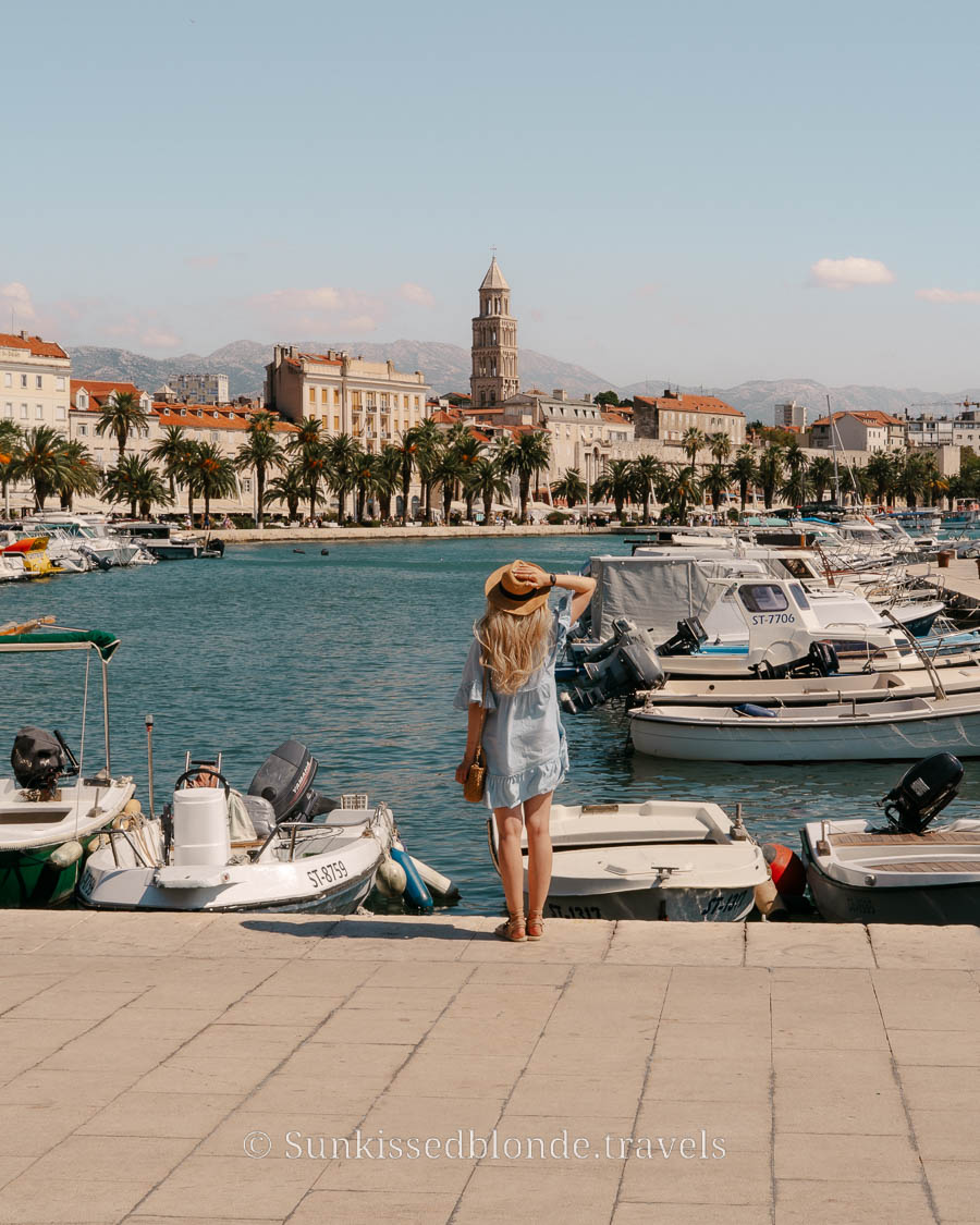 Woman standing on the Split waterfront overlooking the Riva promenade, boats in the marina, and the bell tower of Saint Domnius Cathedral.