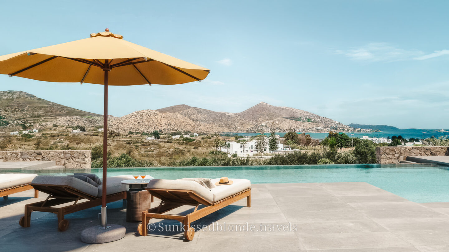 Luxury infinity pool in Paros, Greece, with sun loungers and a yellow umbrella overlooking turquoise water, rolling hills, and white Cycladic houses.