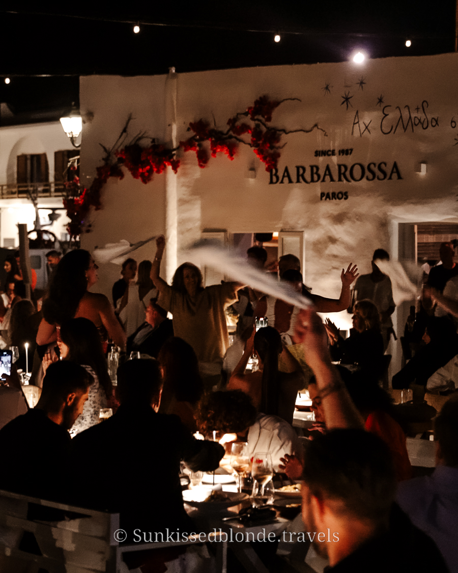Lively evening scene at Barbarossa restaurant in Naoussa, Paros, with crowds dining outdoors, illuminated string lights, and people waving napkins during the traditional celebration.