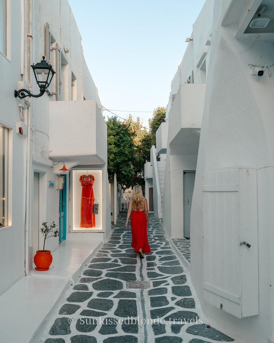 Person walking down a narrow Cycladic alley with whitewashed buildings, stone‑paved paths, and a boutique window displaying a red dress in the Greek islands