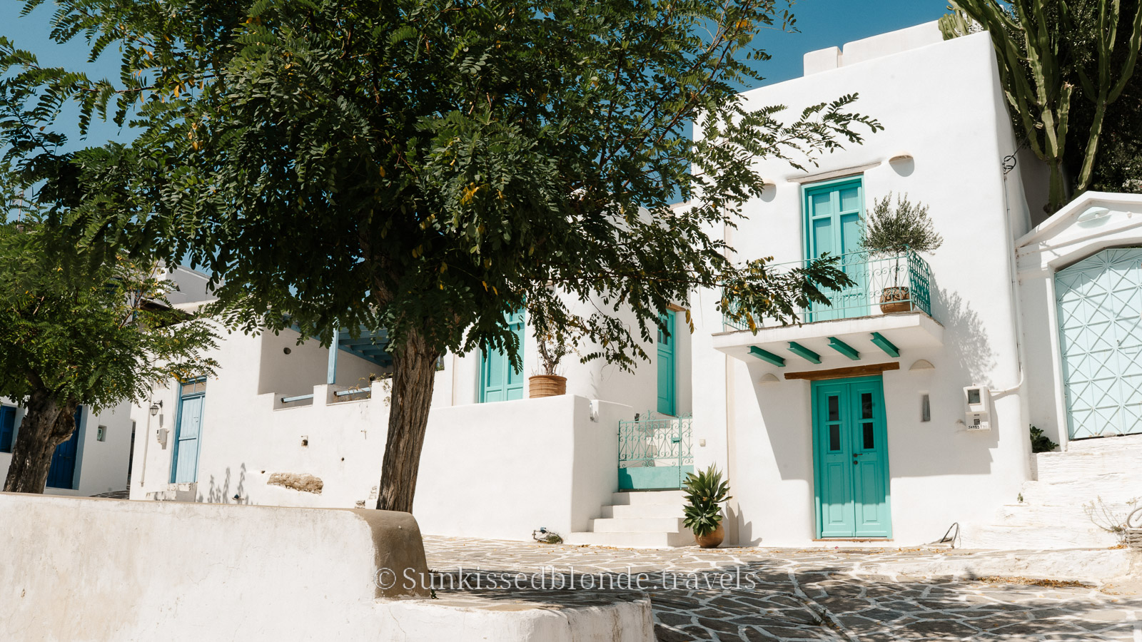 Traditional Cycladic house in Paros, Greece, with whitewashed walls, turquoise doors and windows, shaded by trees along a stone‑paved street
