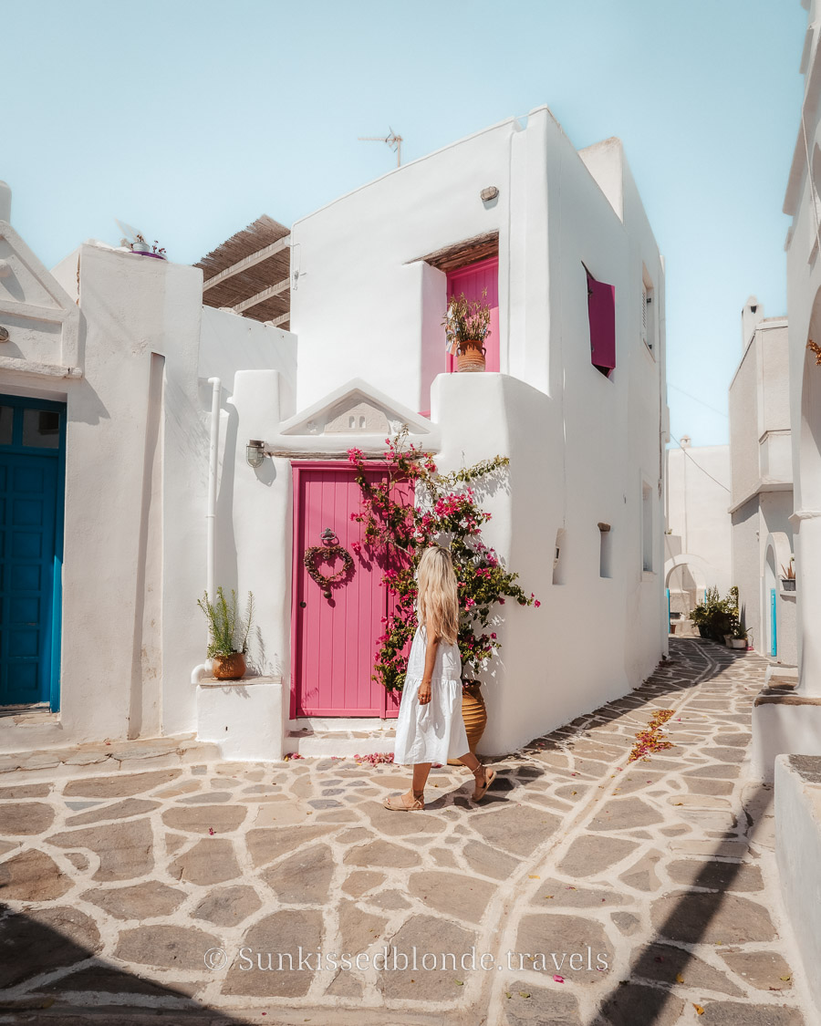 Person walking along a stone‑paved alley in Paros, Greece, past whitewashed Cycladic houses with bright pink doors, shutters, and flowering bougainvillea