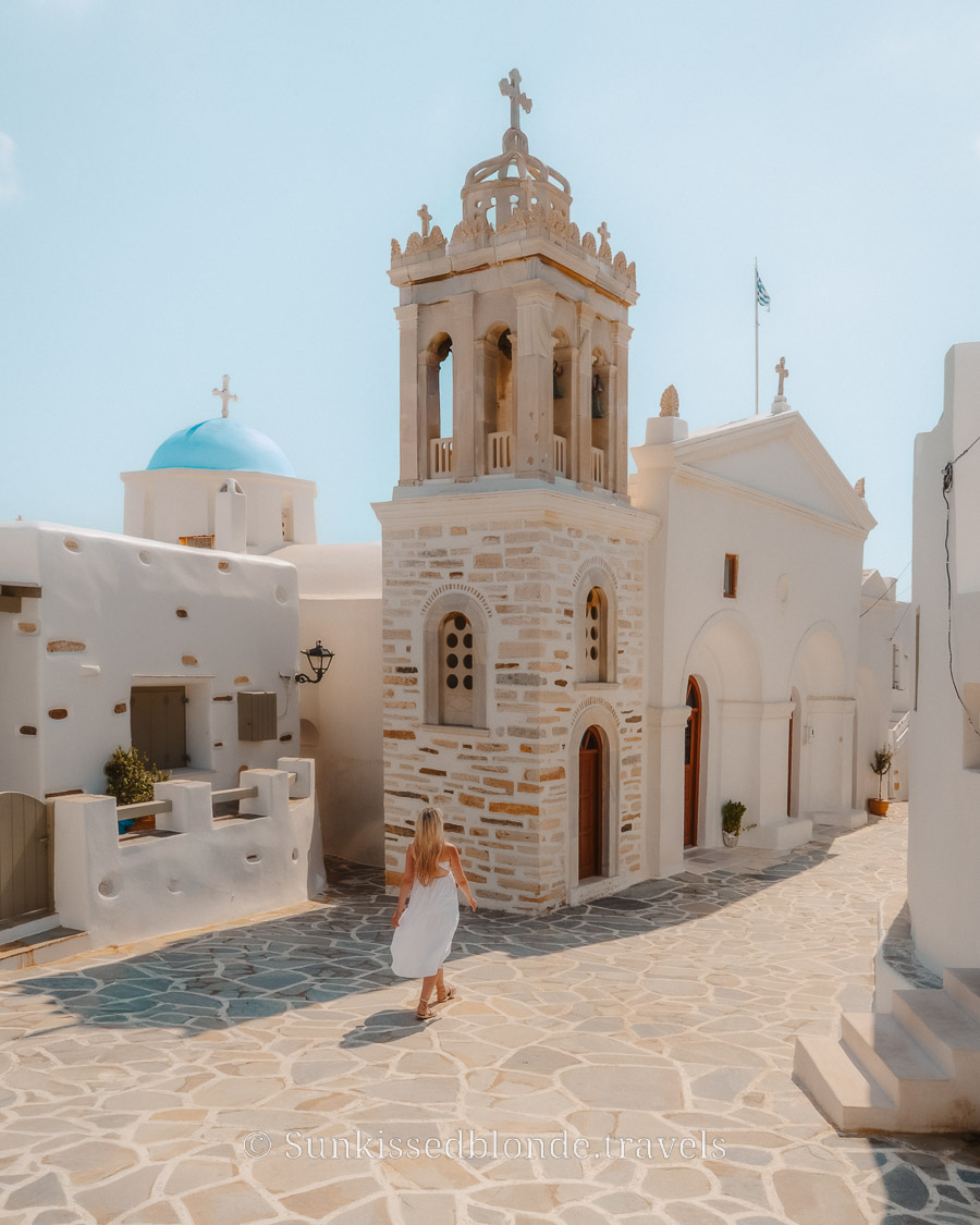 Person walking toward a traditional stone bell tower and whitewashed church in Paros, Greece, surrounded by Cycladic architecture and a blue‑domed chapel.