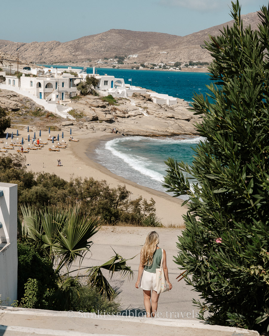 Person walking toward a sandy beach on Paros, Greece, with turquoise water, gentle waves, and white Cycladic buildings perched on rocky cliffs in the distance.