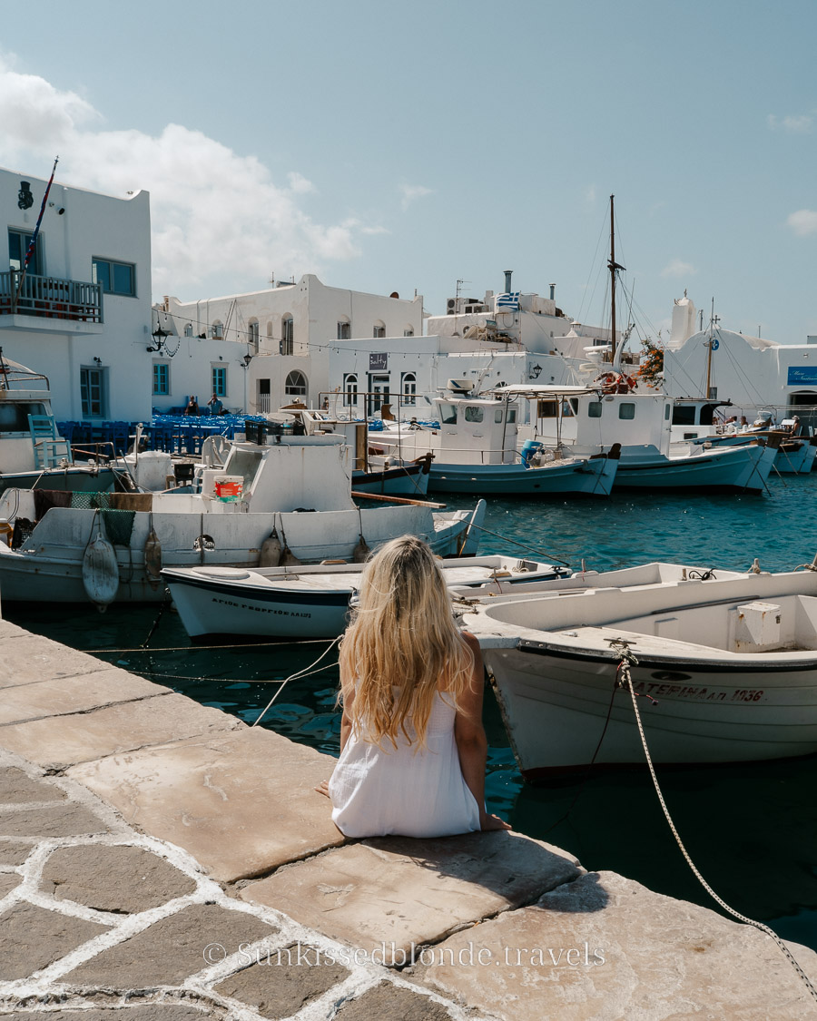 Calm harbour scene in Naoussa, Paros, with a person seated by the waterfront facing moored fishing boats and classic whitewashed architecture.