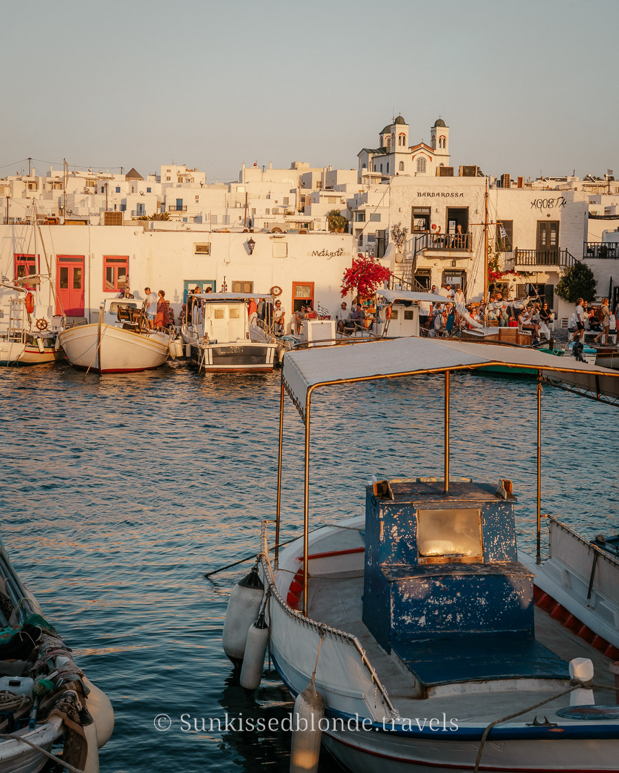 Fishing boats docked in the Naoussa harbour of Paros, Greece, with whitewashed Cycladic buildings and a church with twin bell towers at sunset.
