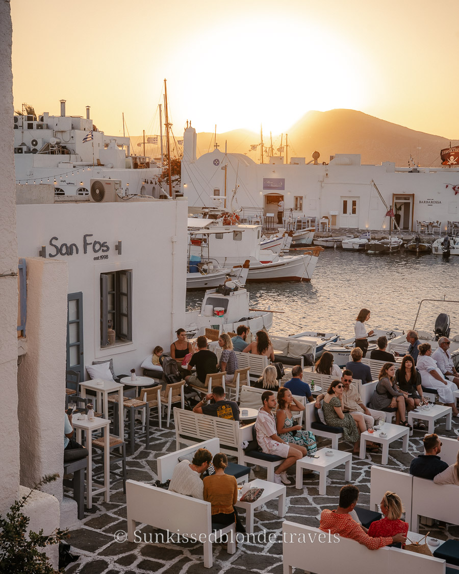 Sunset at the Naoussa harbour in Paros, Greece, with people seated at waterfront caf&eacute;s, white Cycladic buildings, and fishing boats docked along the marina.