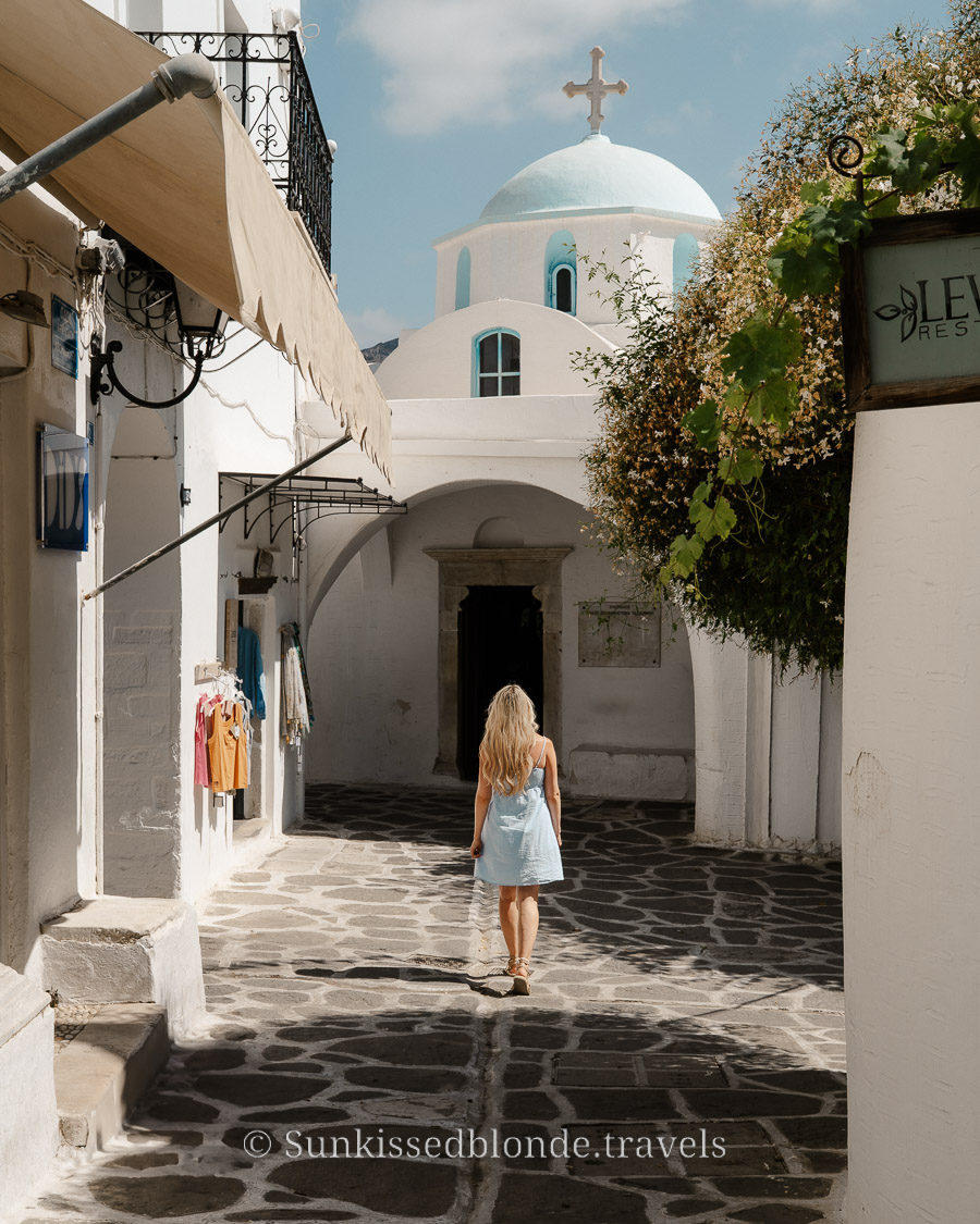 Person walking through a narrow stone‑paved alley in Paros, Greece, surrounded by whitewashed Cycladic buildings and a blue‑domed church with a cross above.
