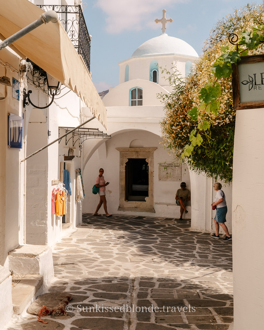 Sunlit alleyway in Paros, Greece, with whitewashed Cycladic buildings, stone‑paved paths, and a domed church with a cross rising above the rooftops.