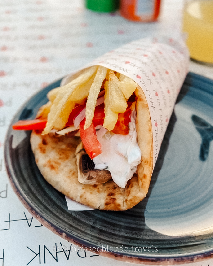 Close‑up of a Greek gyros wrap on a plate, filled with grilled meat, tomatoes, onions, tzatziki, and topped with crispy fries.