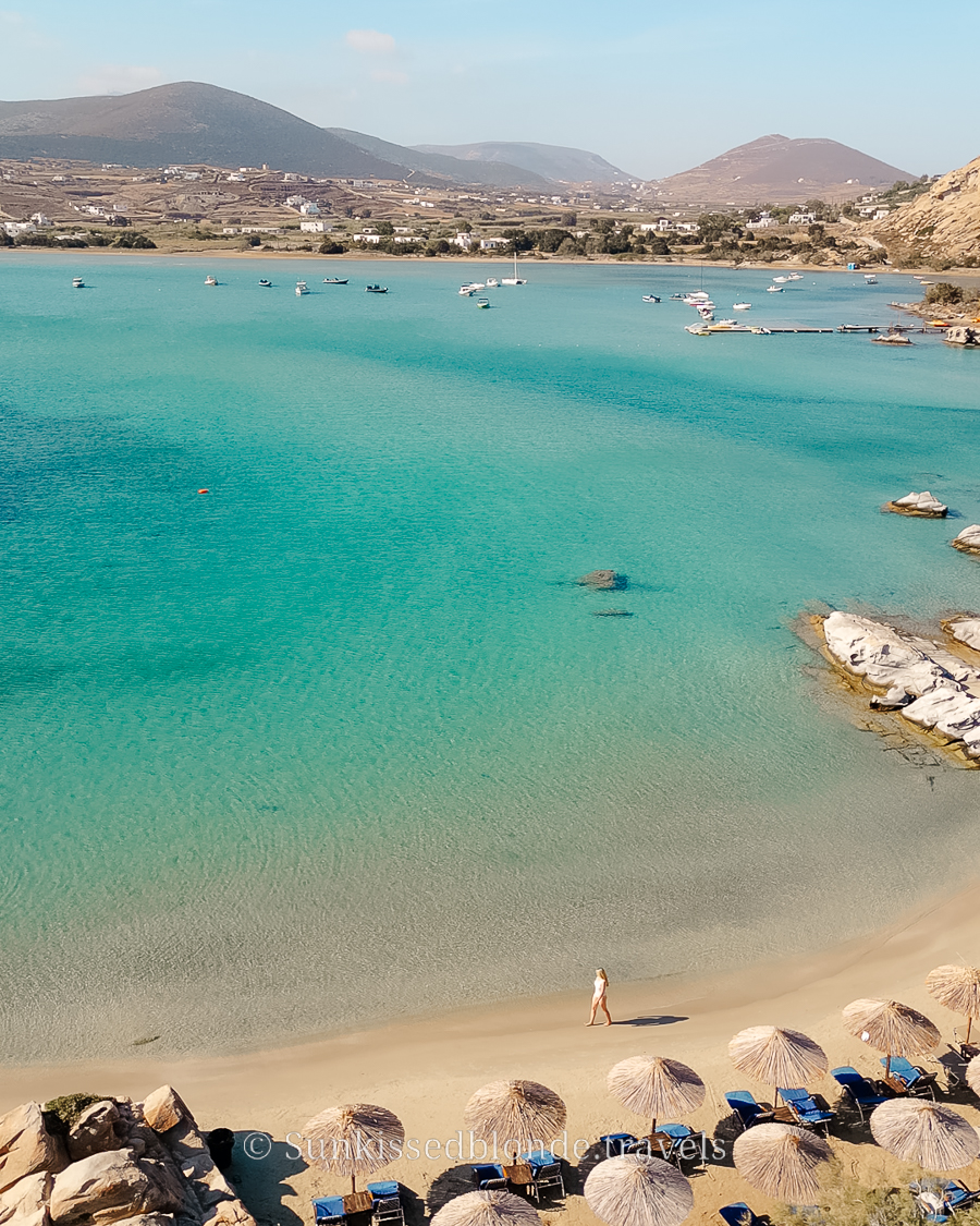 Aerial view of a turquoise bay in Paros, Greece, with clear shallow water, sandy shoreline, sunbeds with straw umbrellas, and a person walking along the beach.
