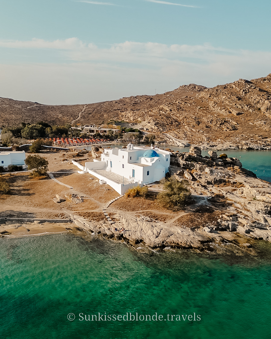 Aerial view of a whitewashed church with a blue dome on the rocky coastline of Paros, Greece, surrounded by turquoise water and rugged hills.