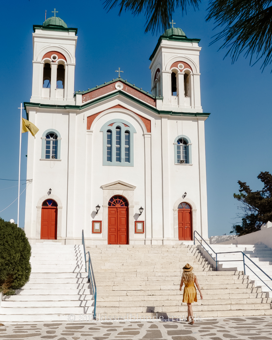Person walking up the stone steps toward a large white Orthodox church in Paros, Greece, featuring twin bell towers, red doors, and green‑trimmed domes under a clear blue sky.