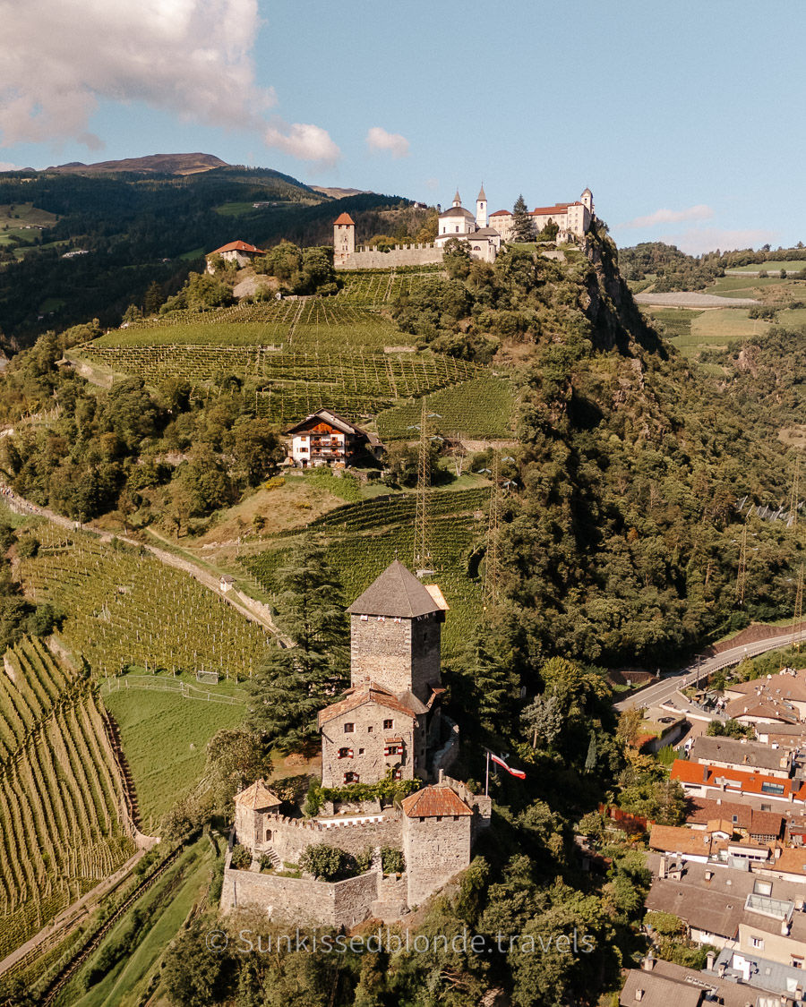 Aerial view of Sabiona Monastery and Branzoll Castle above vineyard-covered hills in Chiusa (Klausen), South Tyrol, surrounded by alpine landscape and village rooftops