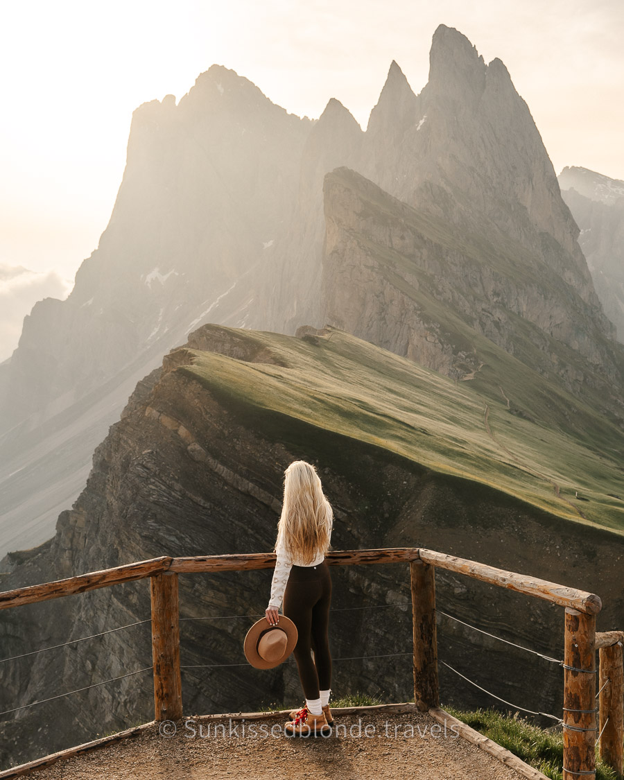 Iconic Seceda ridgeline in the Dolomites featuring layered rock formations, grassy slopes, and misty Sunrise light over the mountains with Laura Sunkissedblonde.travels standing at the viewpoint.