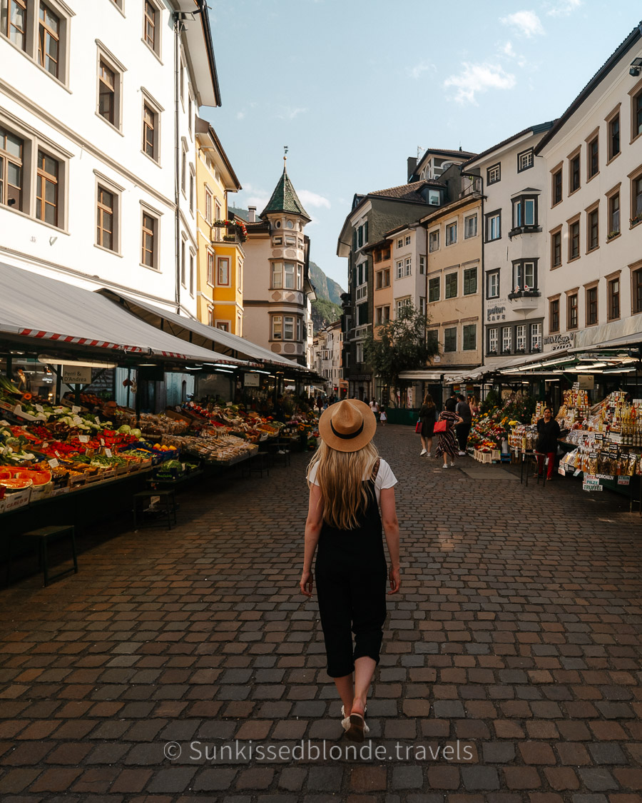 Person walking along the cobblestone market street in Bolzano, South Tyrol, Dolomites surrounded by colorful historic buildings and open‑air stalls selling fresh produce and local goods.