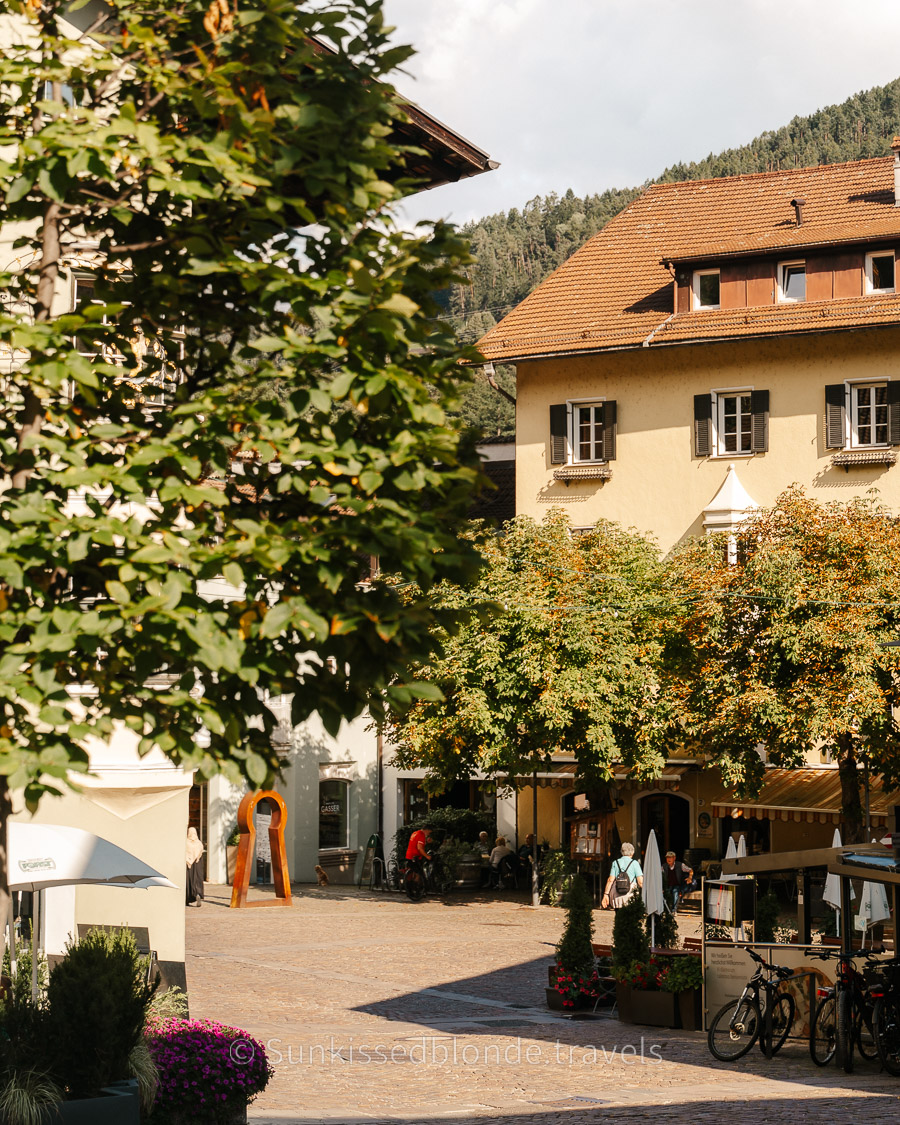 Sunny town square in Chiusa (Klausen), South Tyrol, with leafy trees, outdoor café seating, bicycles, and traditional alpine buildings with shuttered windows.