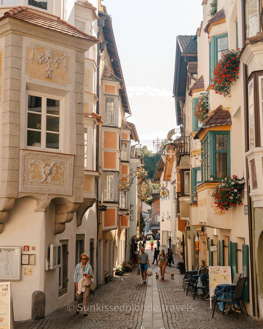 Charming old town street in Chiusa (Klausen), South Tyrol, lined with colorful historic buildings, ornate bay windows, hanging signs, and cobblestone pathways.