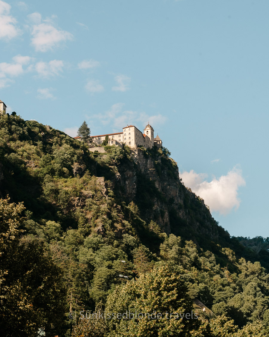 Sabiona Monastery perched on a steep rocky hillside above Chiusa (Klausen) in South Tyrol, surrounded by dense green forest and a clear blue sky