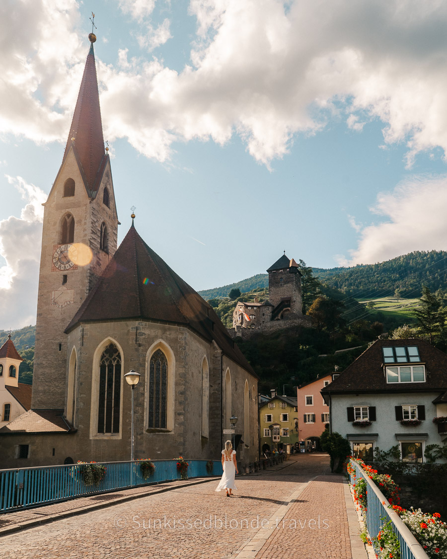Mountain village of Chiusa scene in South Tyrol with a tall Gothic church tower, pastel houses, and a hillside castle above, with a person walking across a flower‑lined bridge