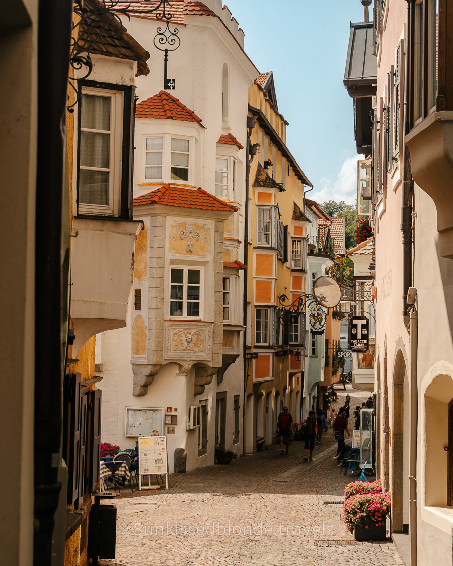 Medieval street in Chiusa (Klausen), South Tyrol, lined with ornate pastel buildings, carved bay windows, wrought‑iron signs, and cobblestone pathways.