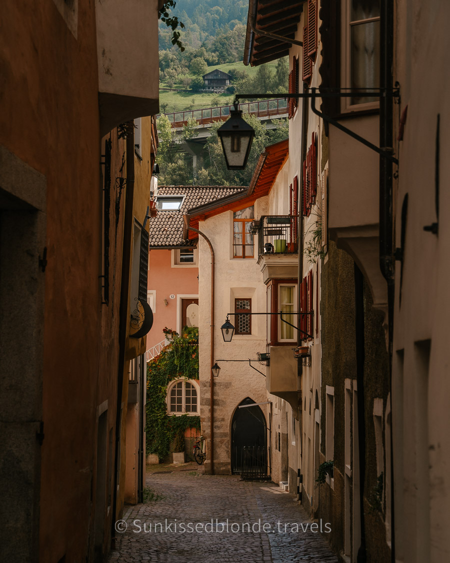 Narrow cobblestone alleyway in Chiusa (Klausen), South Tyrol, lined with pastel buildings, shuttered windows, hanging lanterns, and a view of green hills and a farmhouse in the distance.