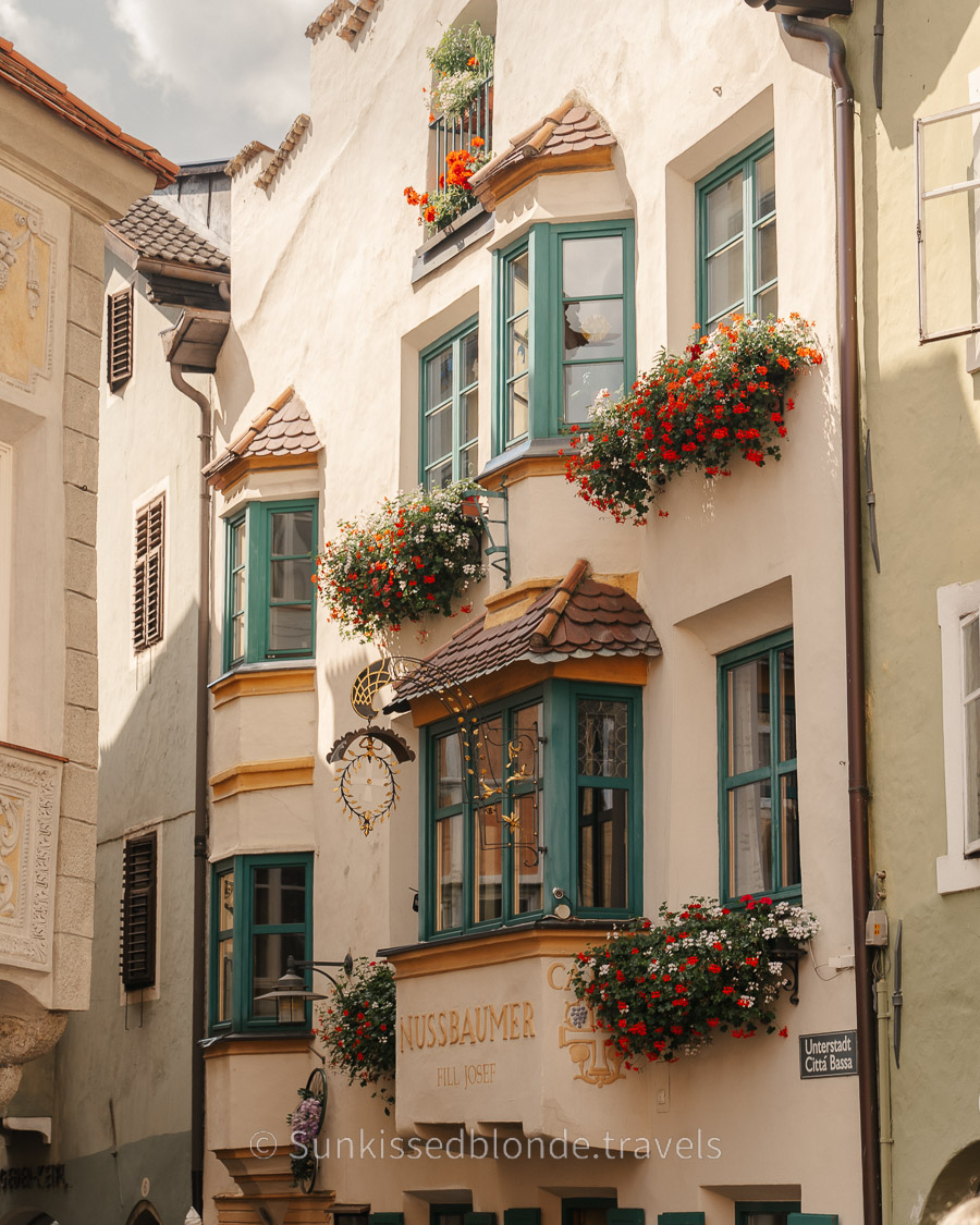 Historic building in Chiusa (Klausen), South Tyrol, featuring ornate bay windows, teal shutters, flower-filled balconies, and traditional Tyrolean architectural details.