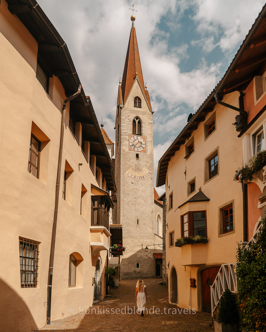 Historic cobblestone street in Chiusa (Klausen), South Tyrol, leading to the tall Gothic bell tower of St. Andrew’s Church, with alpine-style buildings on both sides.