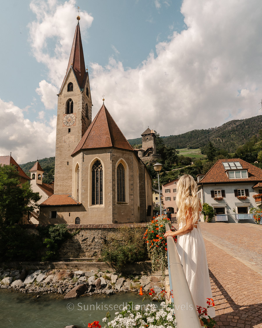 View of the historic church of St. Andrew in Chiusa (Klausen), South Tyrol, with a person standing on a bridge overlooking the river and medieval village framed by mountain scenery.