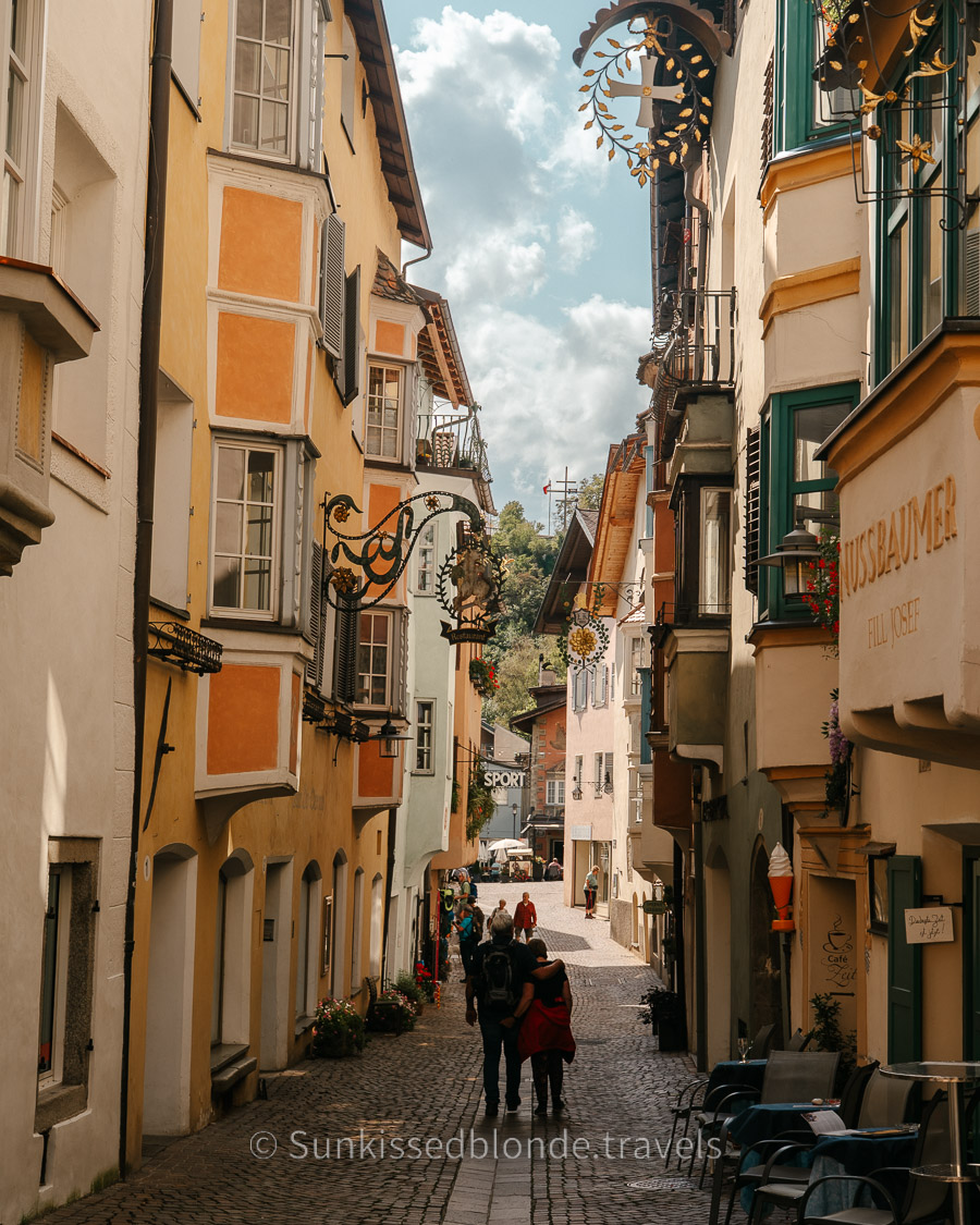 Historic cobblestone street in Chiusa (Klausen), South Tyrol, lined with colorful medieval buildings, ornate wrought‑iron signs, and traditional alpine architectural details