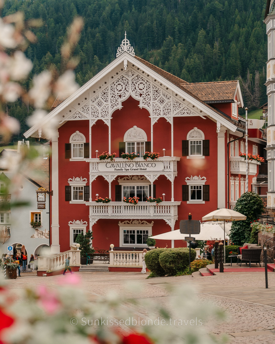 Ornate red alpine building with white decorative woodwork and flower‑filled balconies in the town of Ortisei, Val Gardena in the Dolomites, set against a forested mountain backdrop.