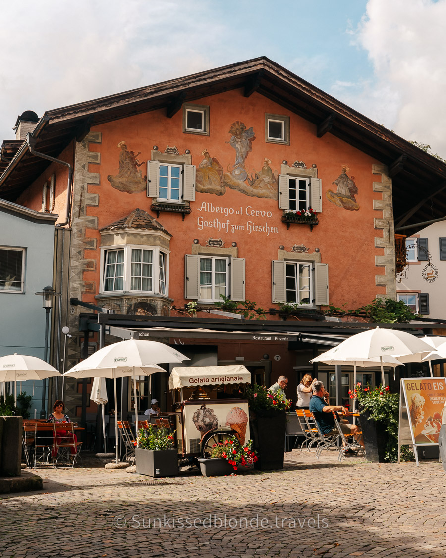 Historic gastropub ‘Albergo al Cervo’ in Chiusa (Klausen), South Tyrol, featuring a painted facade with alpine murals, shuttered windows, and an outdoor terrace with umbrellas and café seating.