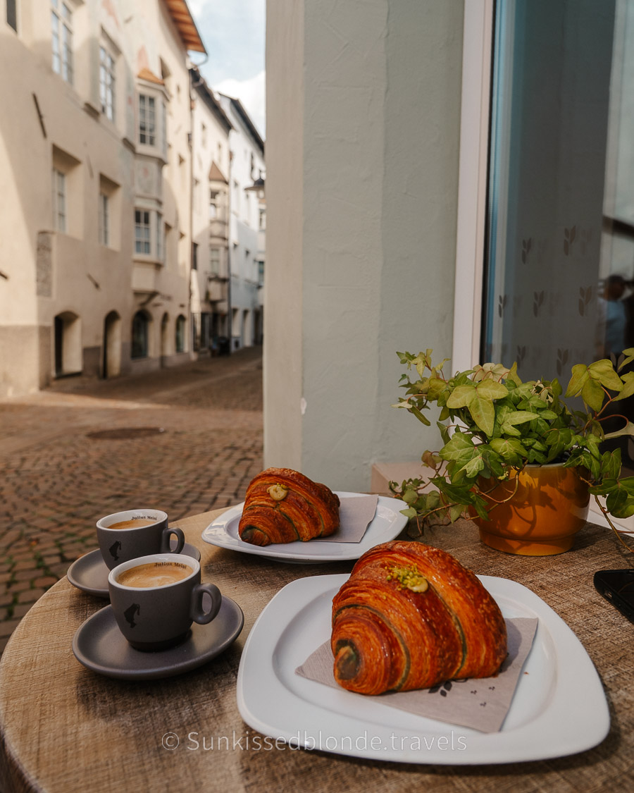 Gasser Bäckerei Outdoor café table in Chiusa (Klausen), South Tyrol, with two pastries and two espresso cups set against a cobblestone street and historic alpine buildings