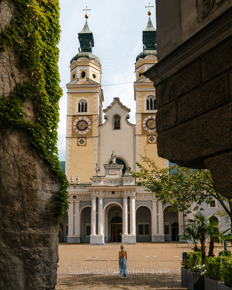 View of Brixen Cathedral in South Tyrol Dolomites framed by stone archways, showing twin bell towers with detailed clock faces and a person walking in the foreground.