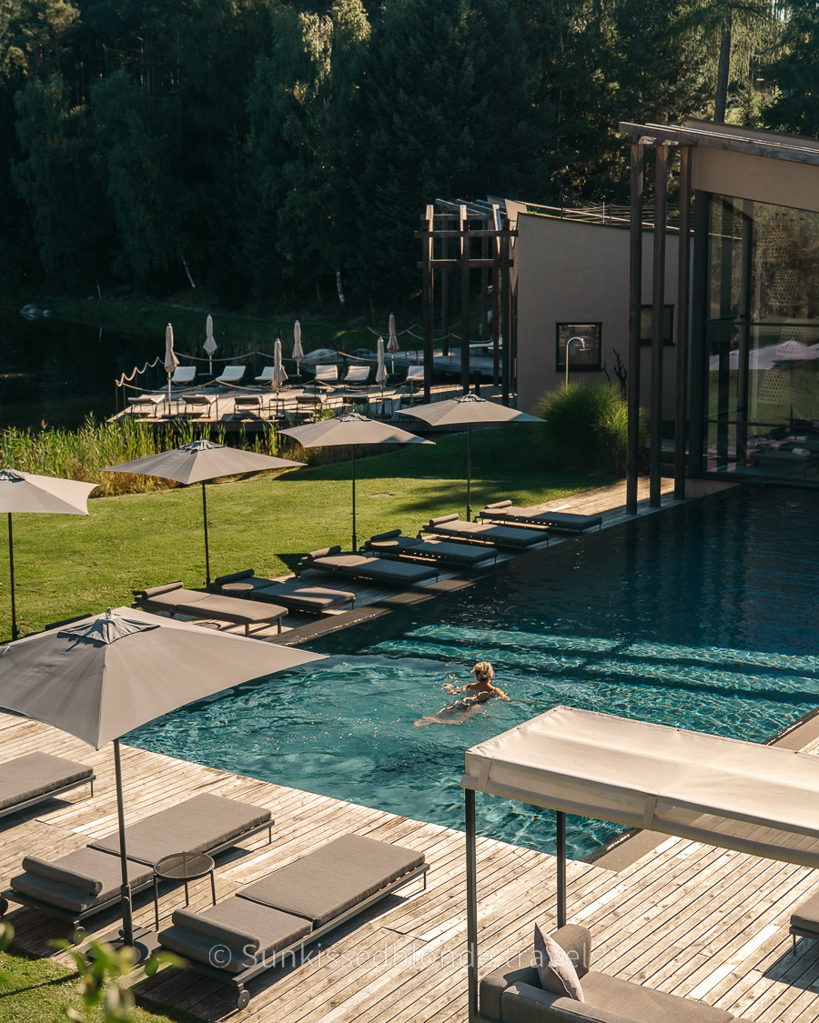 Person swimming in the outdoor infinity pool at Seehof Nature Retreat in South Tyrol, surrounded by forested shoreline and lakeside loungers under umbrellas.