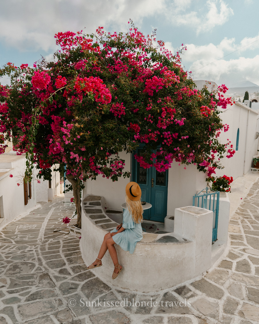 Woman sitting beside a whitewashed house in Paros, Greece, under a vibrant pink bougainvillea tree, surrounded by traditional Cycladic stone paths and blue doors