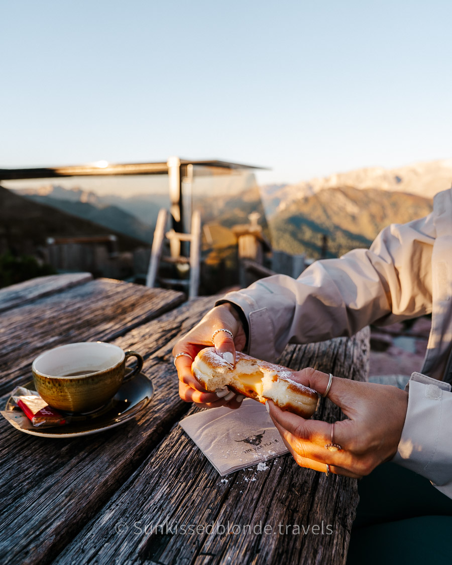 Woman opening doughnut at Rifugio Friedrich August for the Famous Doughnuts at Golden hour light over Alpe di Siusi alpine meadow with Sassolungo mountain peaks in the background, Dolomites, South Tyrol, Italy