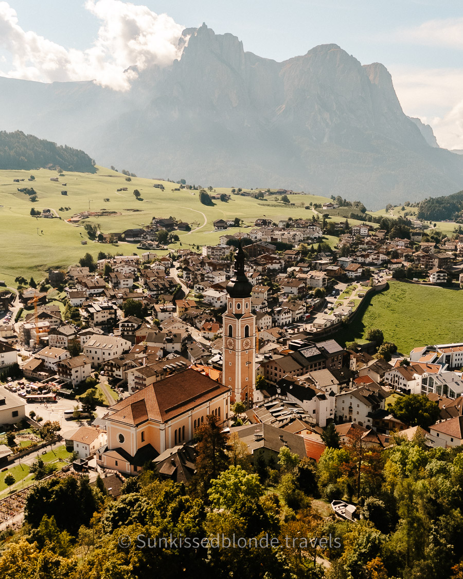 Bird's eye view of Castelrotto (Kastelruth) nestled among alpine meadows with the Dolomite mountains rising beyond, Seiser Alm region, Italy